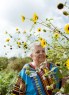 An older pastor stands among tall yellow sunflowers, wearing a brightly patterned blue top.