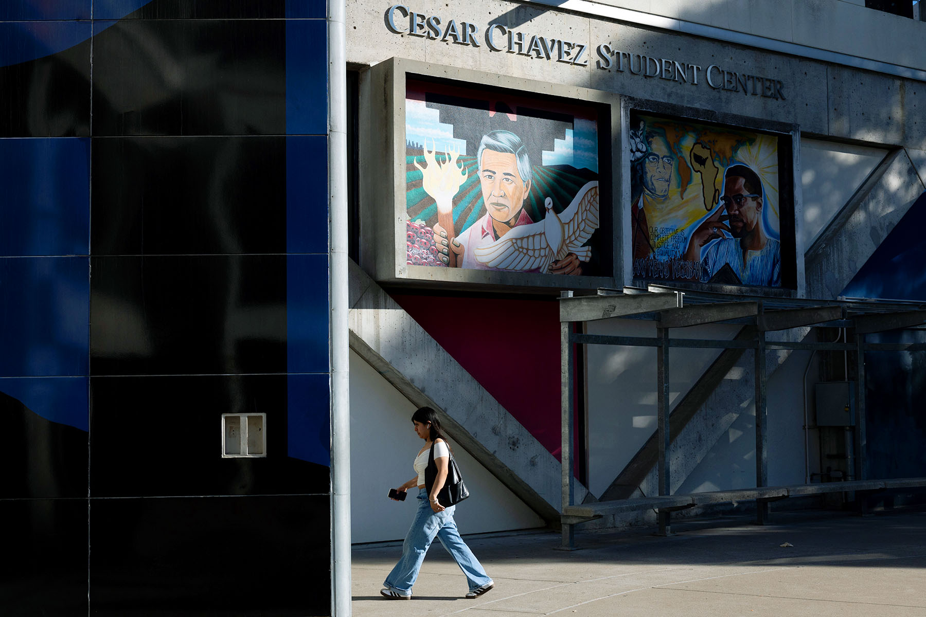 A person walks past a building with a sign reading “Cesar Chavez Student Center,” with painted murals of Cesar Chavez and Malcolm X on the wall above.