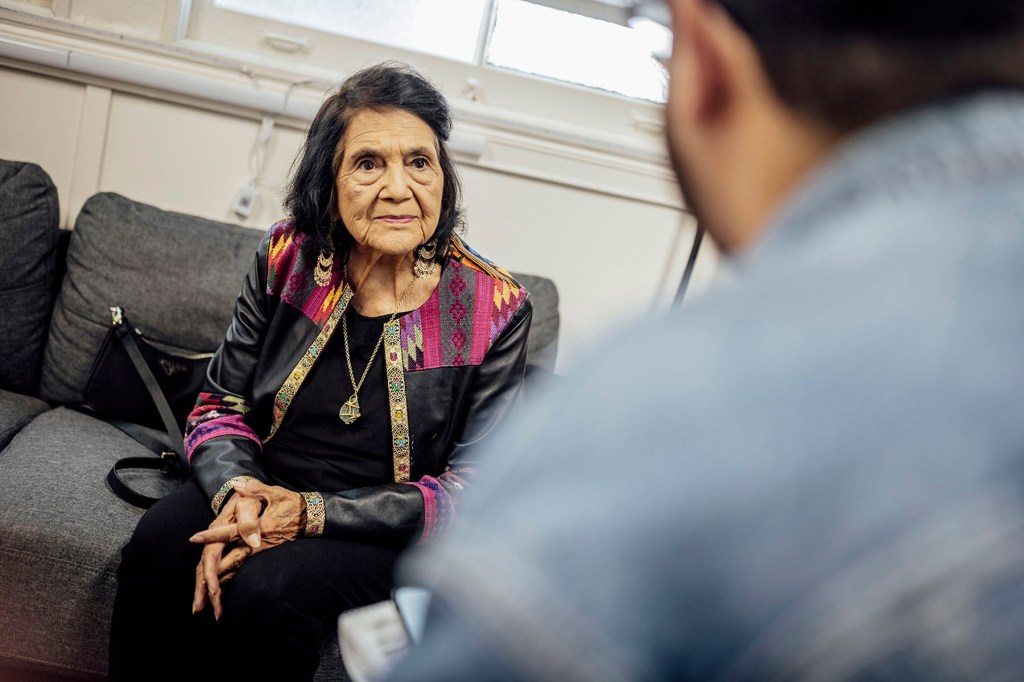 An older woman sits on a couch speaking to someone out of frame, wearing a black outfit with a colorful patterned jacket and gold jewelry, hands clasped as she listens intently.