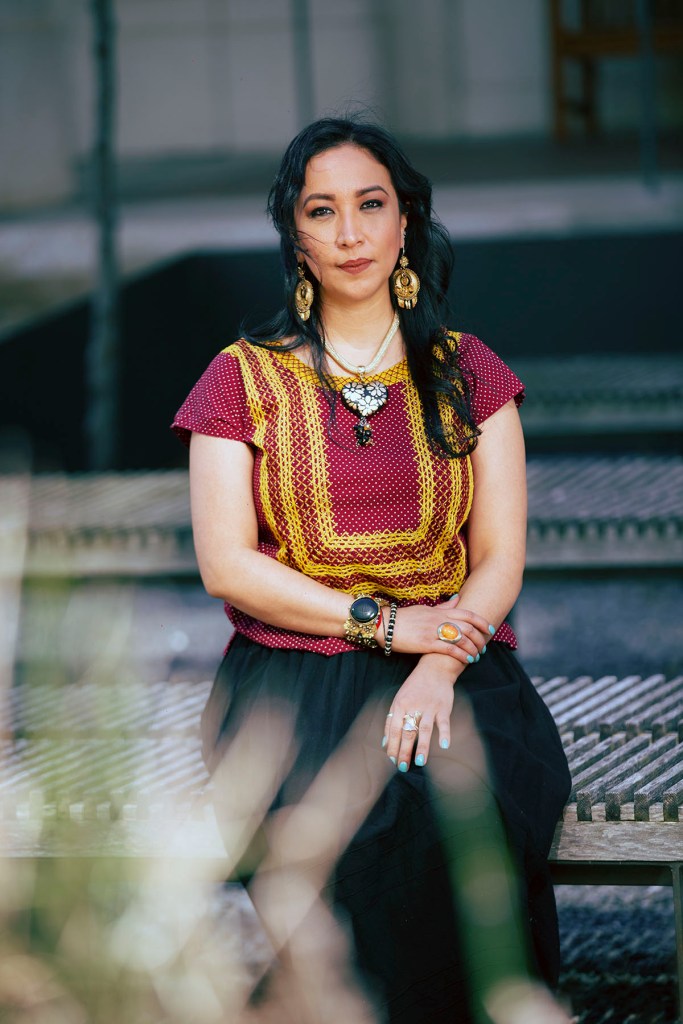A woman with long dark hair sits outdoors on a bench wearing a red and yellow patterned top and black skirt, looking directly at the camera with a composed expression.