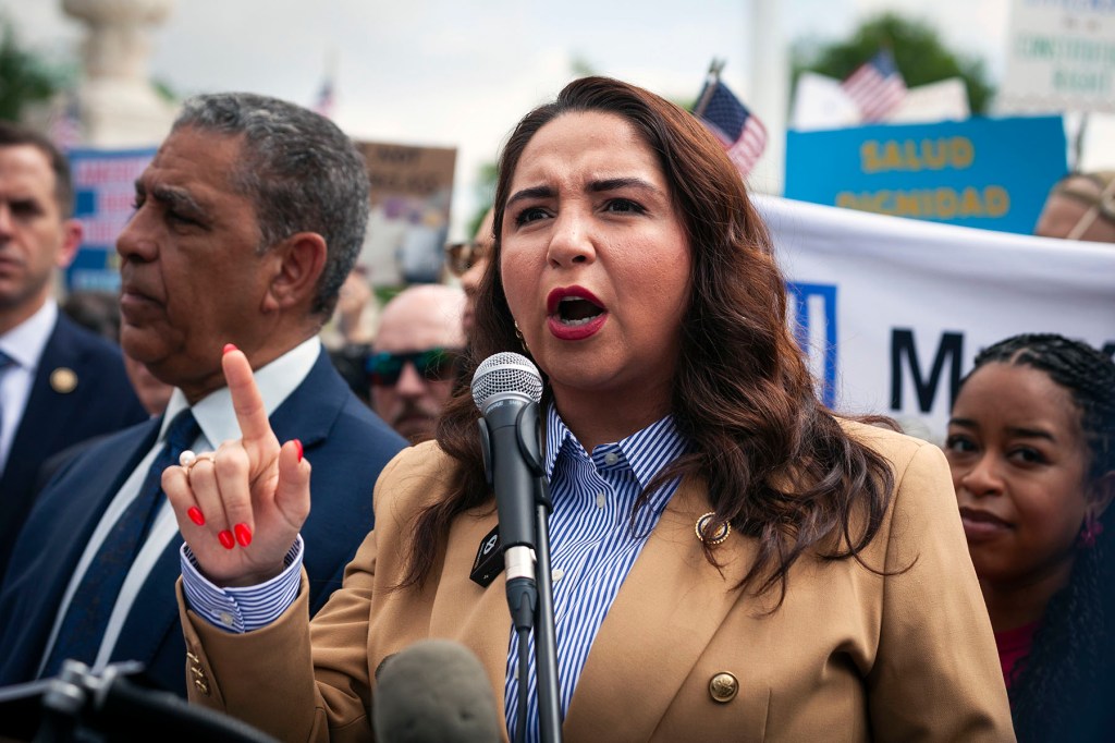A woman speaks into a microphone at a rally, raising one finger as she addresses a crowd with signs and people behind her.