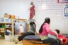A pregnant woman sits on a padded bench, looking down at her belly, while another woman holds a baby nearby in a room with religious imagery, including a statue and portrait of Jesus, and posters on the wall.