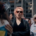 A young woman with pink hair chants during a protest, surrounded by other women.