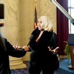 A woman with blonde hair speaks to reporters inside a marble-walled government building, gesturing as a microphone is held toward her. A U.S. flag and a podium with the Senate seal are visible behind her, with cameras and boom microphones framing the scene.