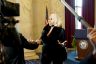 A woman with blonde hair speaks to reporters inside a marble-walled government building, gesturing as a microphone is held toward her. A U.S. flag and a podium with the Senate seal are visible behind her, with cameras and boom microphones framing the scene.