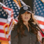 Noem stands in front of several American flags in militaristic attire.