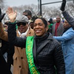 Lt. Governor Juliana Stratton wearing a sash waves to a crowd while walking in a St. Patrick's Day parade, surrounded by people dressed in green and winter clothing.