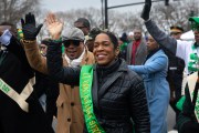 Lt. Governor Juliana Stratton wearing a sash waves to a crowd while walking in a St. Patrick's Day parade, surrounded by people dressed in green and winter clothing.