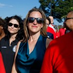 Jocelyn Benson wearing sunglasses stands in a crowd at an outdoor event, with people around her in bright daylight.