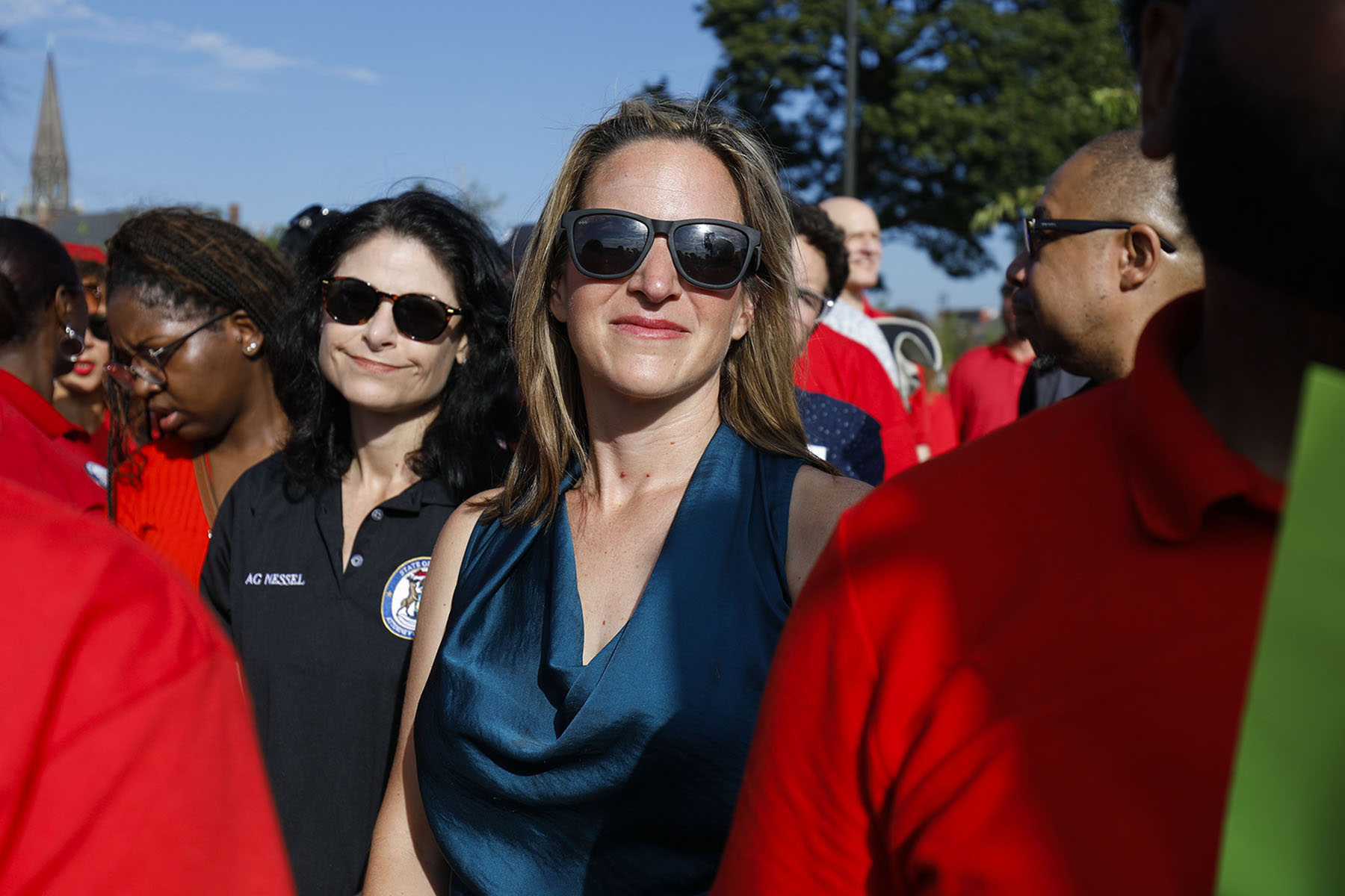 Jocelyn Benson wearing sunglasses stands in a crowd at an outdoor event, with people around her in bright daylight.