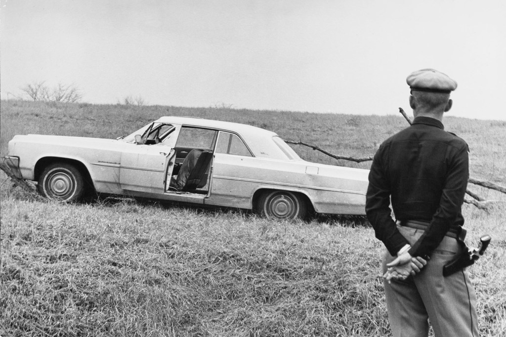 Black and white image of a car stopped on a grassy roadside with its door open, as a state trooper stands nearby observing the scene.