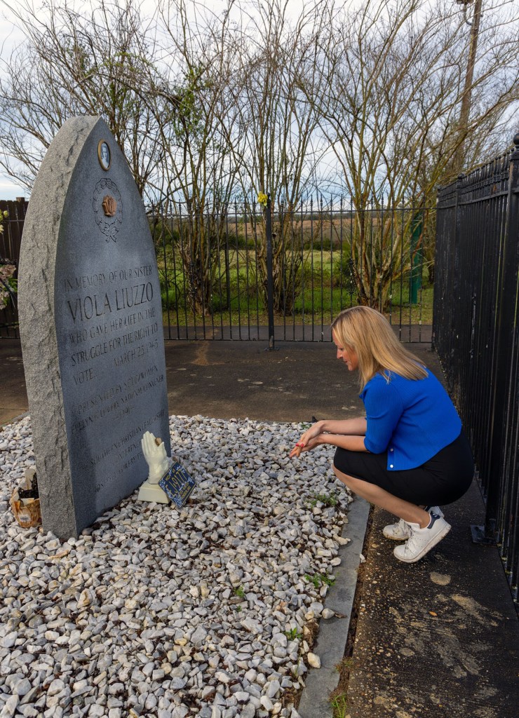 Jocelyn Benson crouches beside a gravestone memorial for Viola Liuzzo, looking down at items placed at its base.