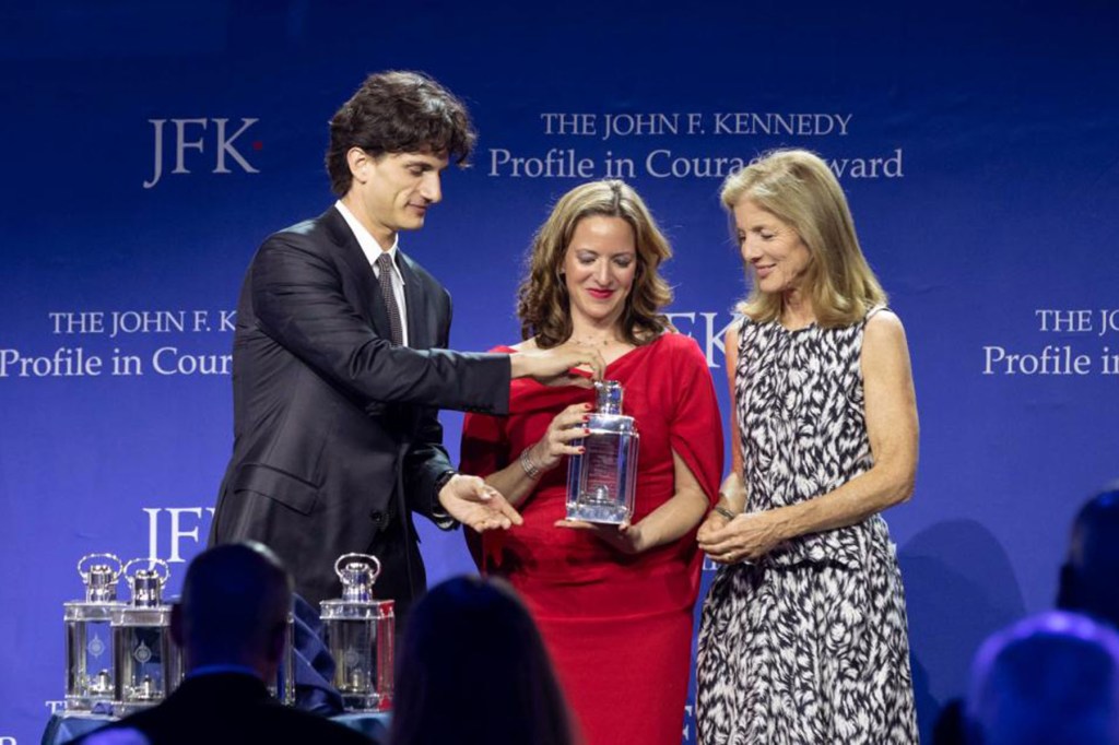 Three people stand on a stage as Jocelyn Benson accepts an award, with a blue backdrop displaying text about the Profile in Courage Award.