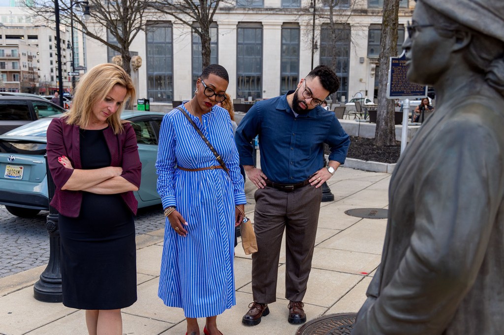 Three people stand on a sidewalk looking toward a statue, appearing reflective as they observe it together.