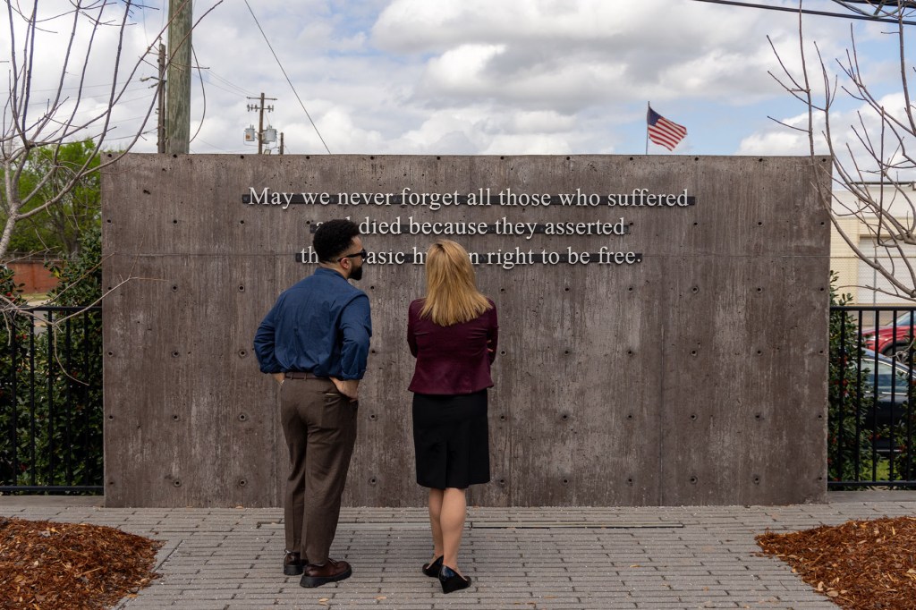 Two people stand facing a large stone wall engraved with text at an outdoor memorial site.