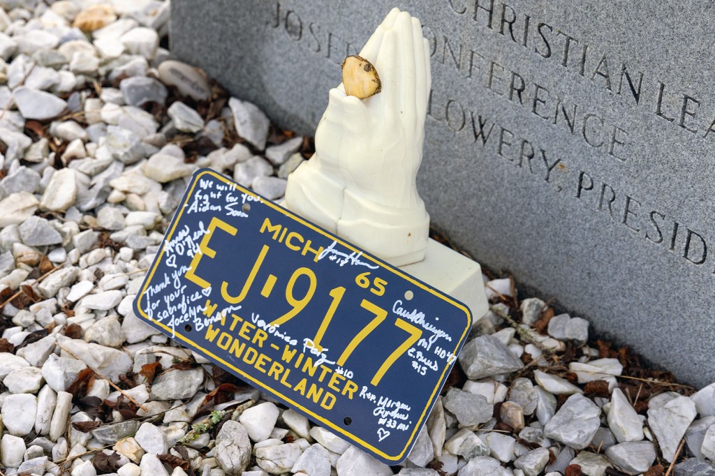 Close-up of a blue Michigan license plate with signatures, placed beside a small memorial object near a gravestone.