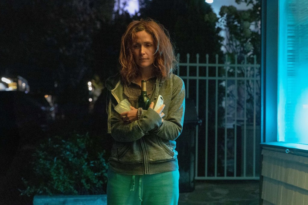 Rose Byrne stands outside at night holding a bottle of wine and a phone, looking tired and contemplative.