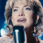 Kate Hudson performs on stage as Claire “Thunder” Sardina, singing into a microphone under bright stage lights.
