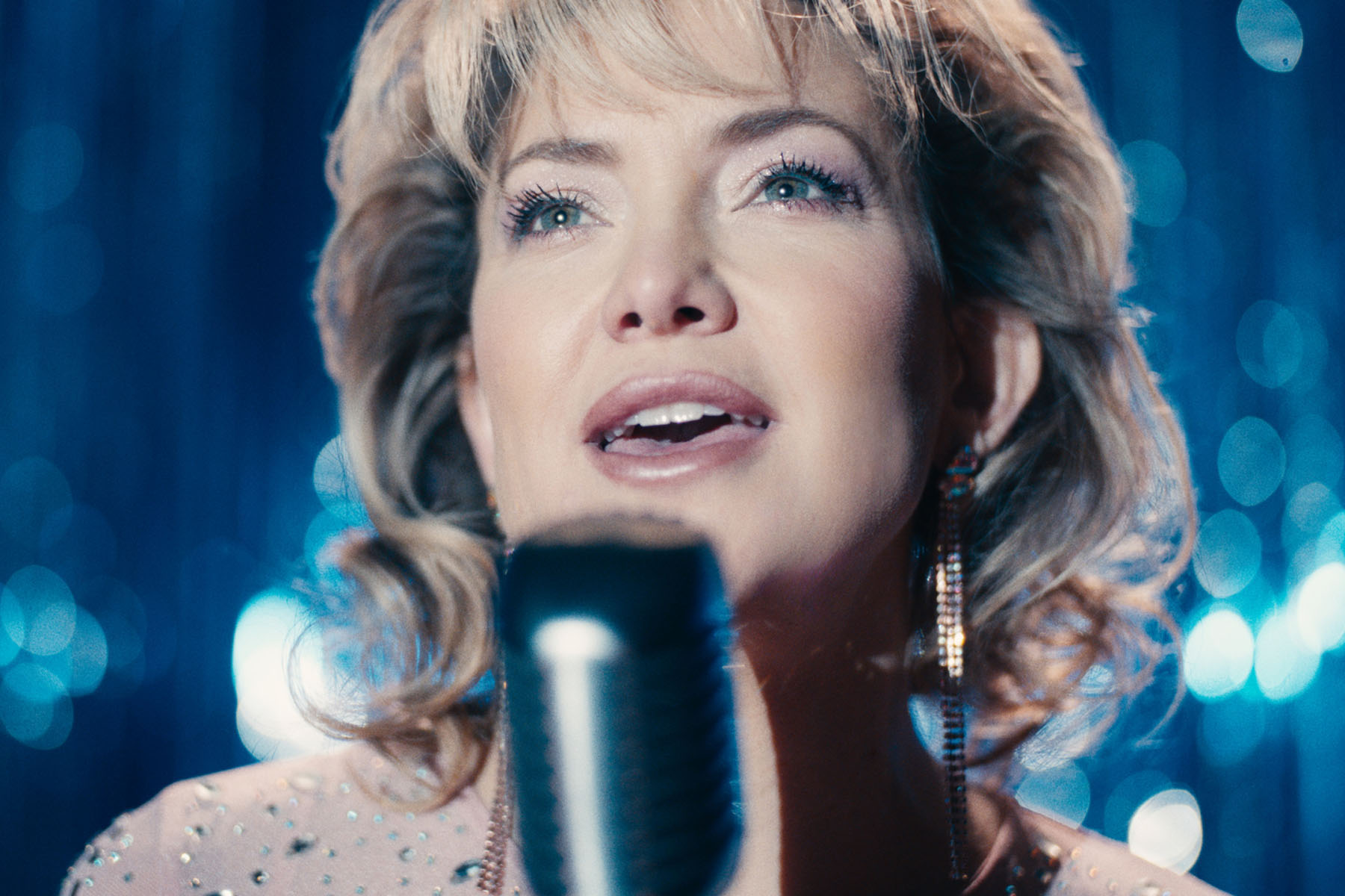 Kate Hudson performs on stage as Claire “Thunder” Sardina, singing into a microphone under bright stage lights.
