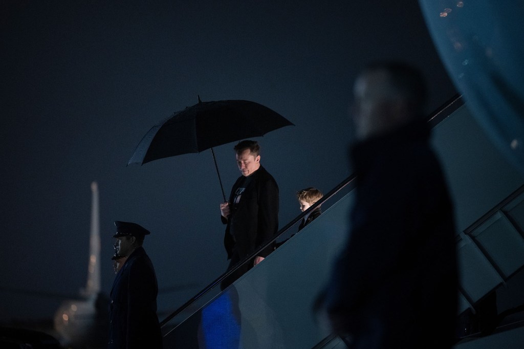 A man holding an umbrella walks down airplane stairs at night, followed by a child, while a uniformed officer stands nearby on the tarmac.