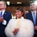 A woman in a white fur wrap smiles and claps while standing between two men in suits at a formal indoor event.