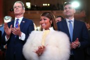 A woman in a white fur wrap smiles and claps while standing between two men in suits at a formal indoor event.