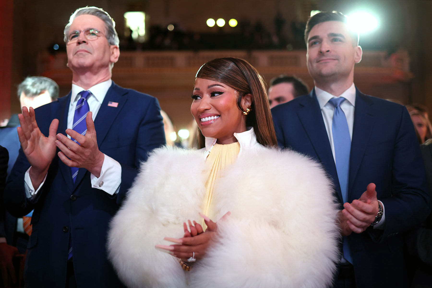 A woman in a white fur wrap smiles and claps while standing between two men in suits at a formal indoor event.