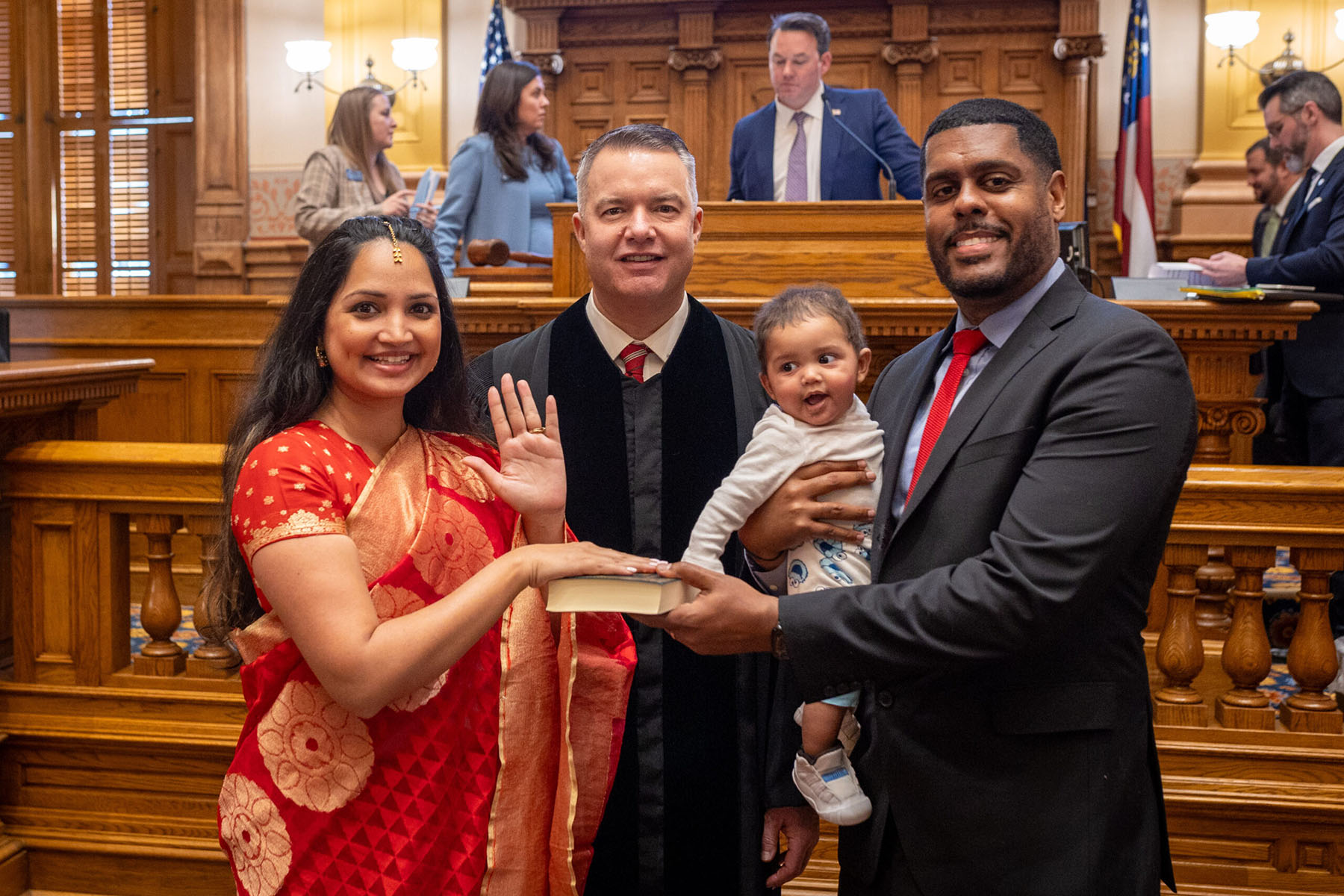 Nabilah Parkes takes the oath of office inside the Georgia Senate chamber, raising her right hand while holding a book, with her husband holding their baby beside her.