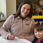 A woman with a child smiles at the camera while signing paperwork.