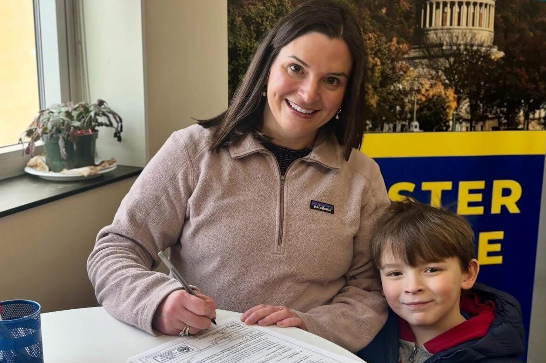 A woman with a child smiles at the camera while signing paperwork.