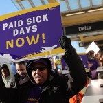 A demonstrator holds a purple and yellow “Paid Sick Days Now!” sign above their head during a rally inside an airport terminal, with other protesters and airline signage visible behind them.