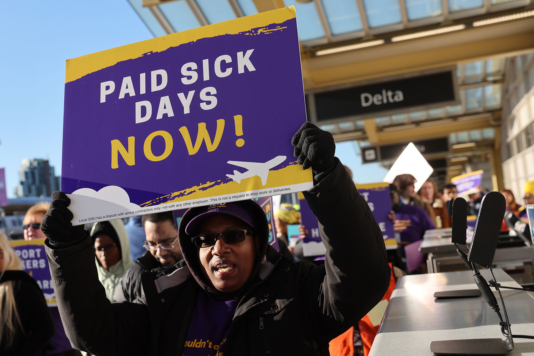 A demonstrator holds a purple and yellow “Paid Sick Days Now!” sign above their head during a rally inside an airport terminal, with other protesters and airline signage visible behind them.