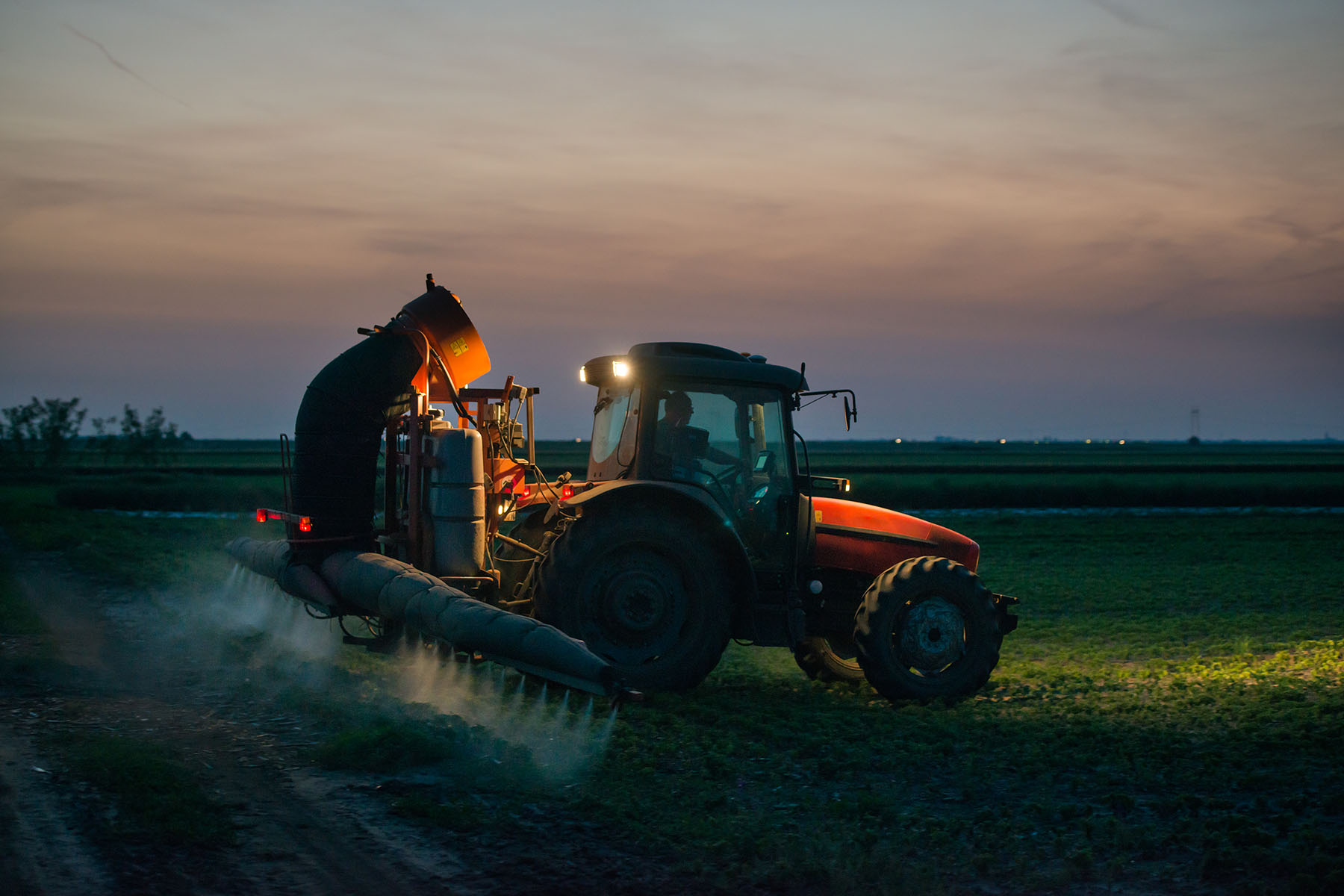 A tractor sprays pesticides crops in a field at sunset.