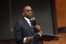 Rev. Jamal Harrison Bryant stands at a podium holding a microphone while speaking during a town hall meeting.