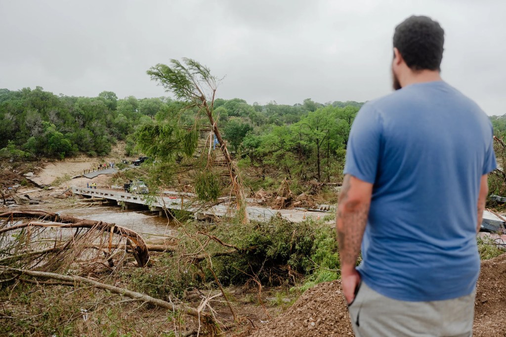 A man stands above a flood-damaged landscape, looking out over a bridge, muddy water, uprooted trees and debris under a gray sky.