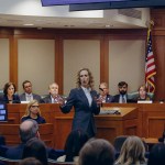 A woman speaks from the center of a committee hearing room, gesturing with both hands as officials sit behind her and audience members watch from the foreground.