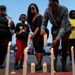 People gather around a row of lit candles at an evening vigil in Shreveport. Several adults and a child lean down to place candles, while others stand nearby in silence.