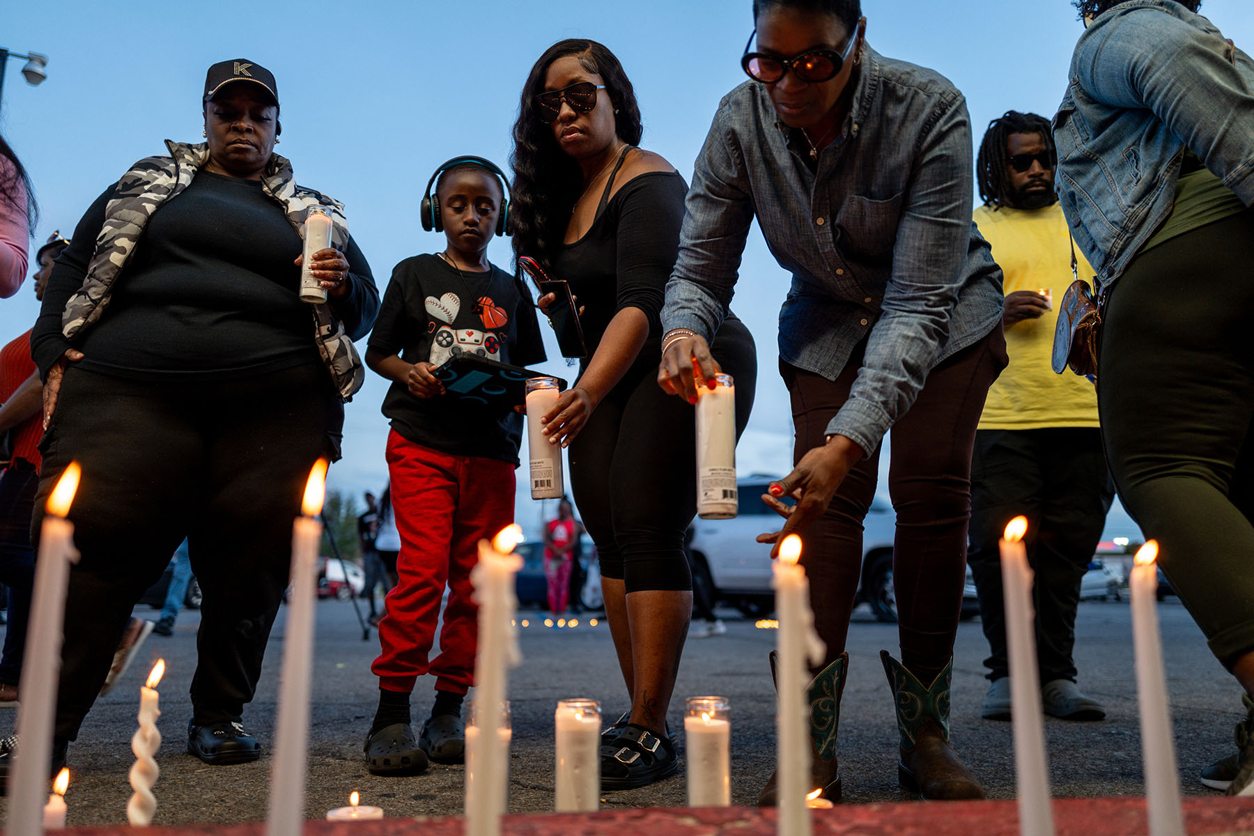 People gather around a row of lit candles at an evening vigil in Shreveport. Several adults and a child lean down to place candles, while others stand nearby in silence.