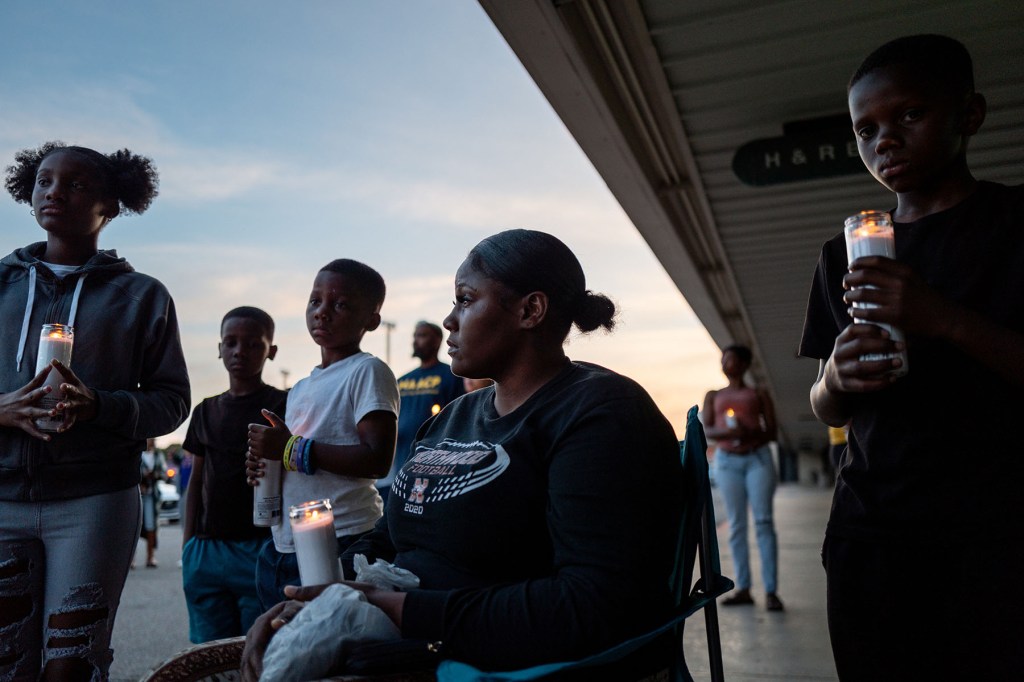 A seated woman and several children hold candles during a vigil at dusk in Shreveport. More people stand in the background outside a strip mall as the sky darkens.