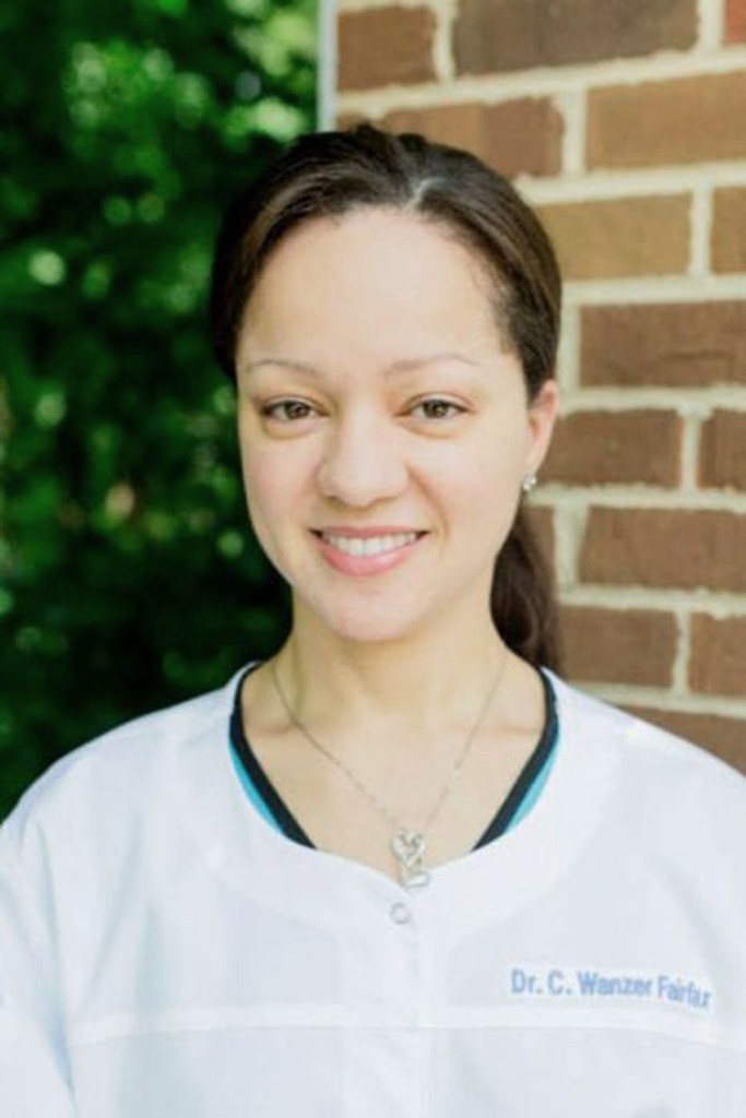 Portrait of Dr. Cerina Fairfax smiling in light-colored medical scrubs, standing in front of a brick wall with green foliage in the background.