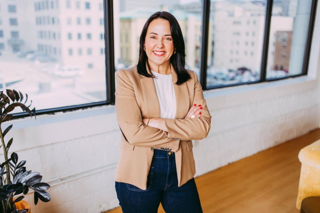 Denise Powell smiles for a portrait with her arms crossed. She is wearing a blazer and standing in front of large windows.