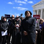 A woman speaks at microphones outside the Supreme Court, surrounded by demonstrators holding signs that read “Haiti is unsafe” and “Protect life — uphold TPS.”