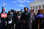 A woman speaks at microphones outside the Supreme Court, surrounded by demonstrators holding signs that read “Haiti is unsafe” and “Protect life — uphold TPS.”