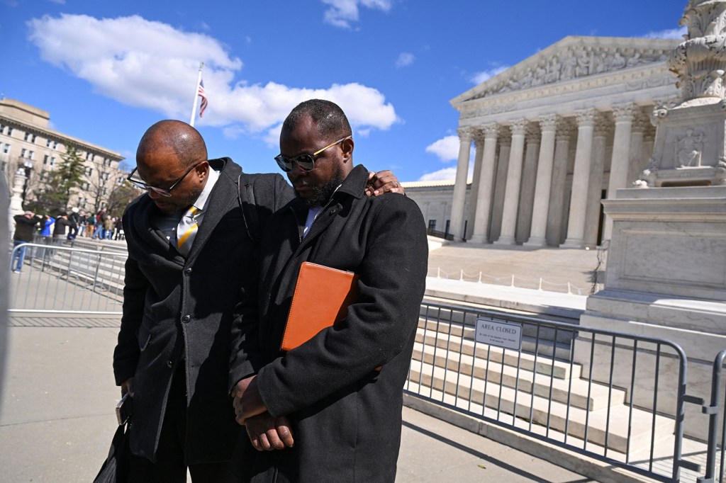 Two men stand with their heads bowed outside the Supreme Court, one with his arm around the other’s shoulder.