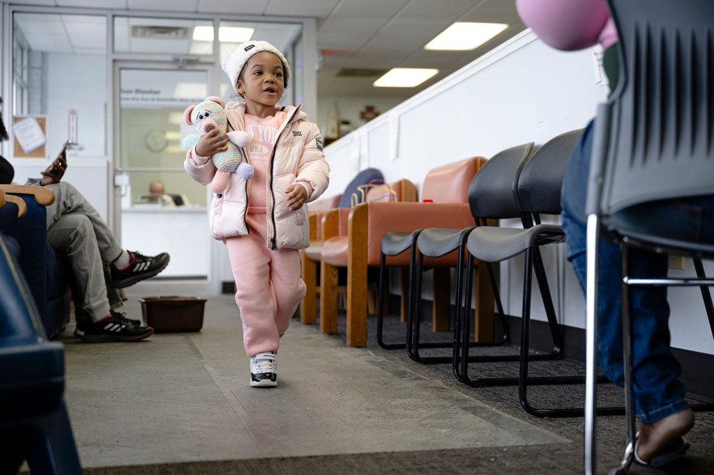 A young child in a pink outfit walks through a waiting room while holding a stuffed animal.