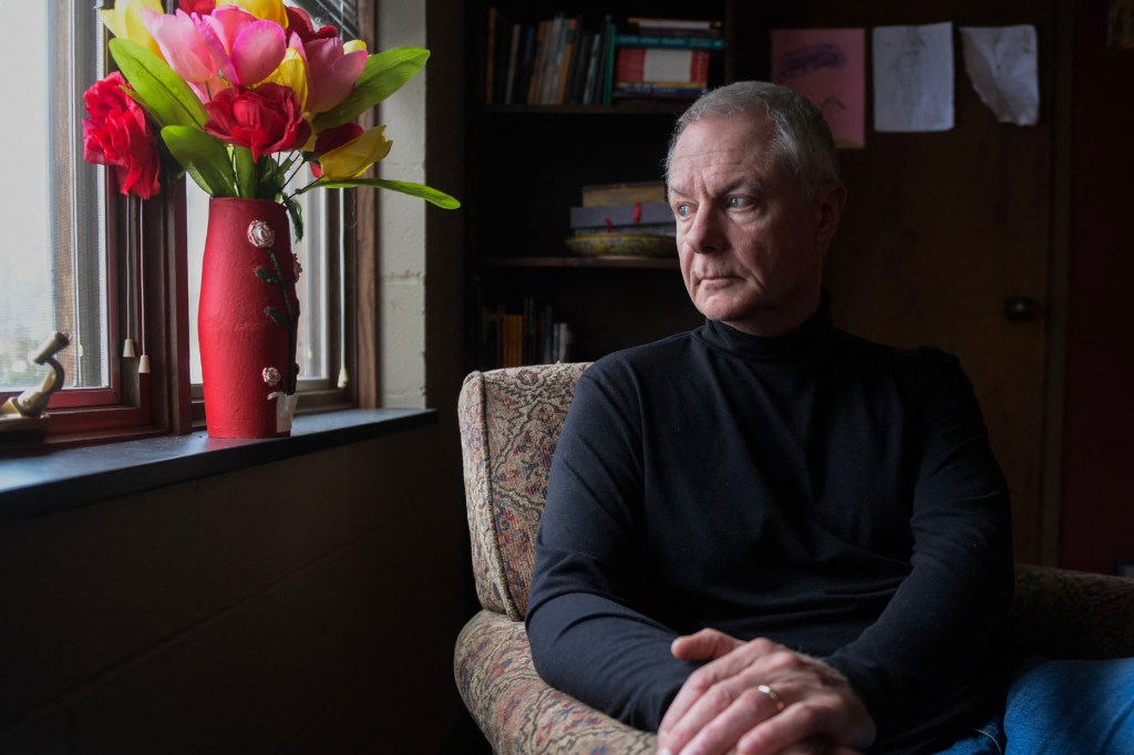 A man in a black turtleneck sits in an armchair beside a window, looking off to the side.