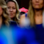 Several women are out of focus in the foreground while a red Make America Healthy Again hat can be seen in the background.