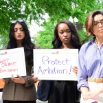 Three women hold signs and stand together at a press conference.