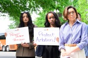Three women hold signs and stand together at a press conference.