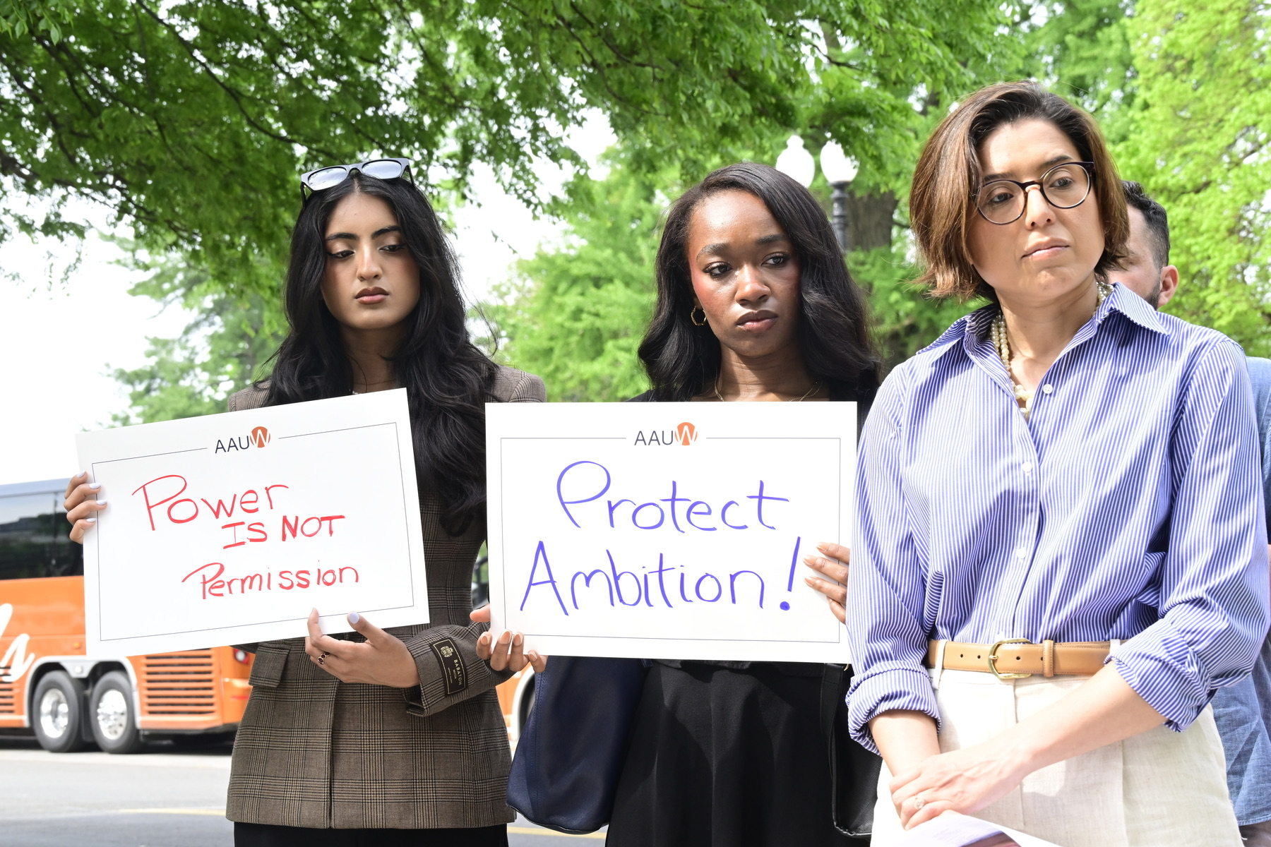Three women hold signs and stand together at a press conference.